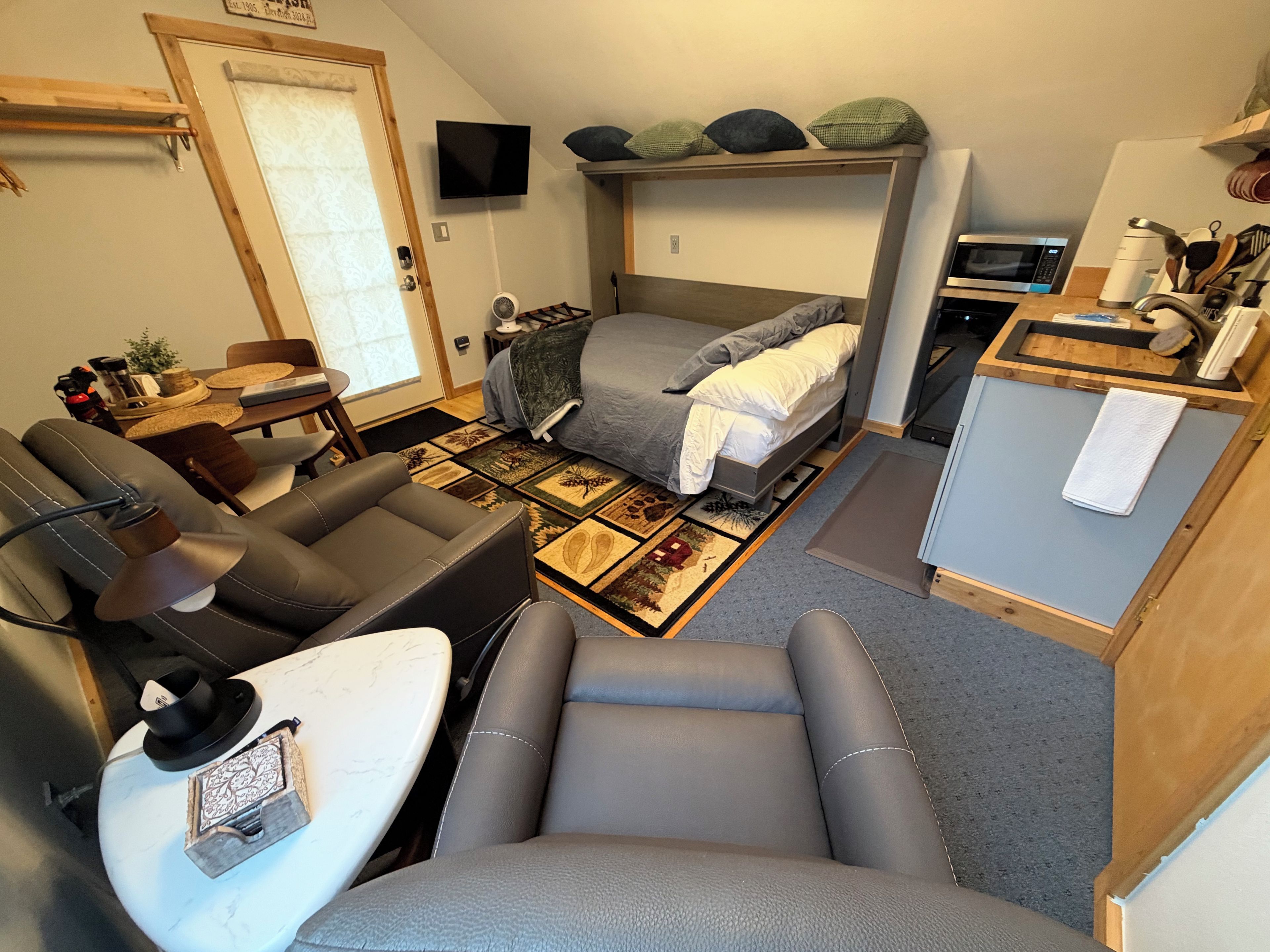 Un espace de vie compact comprenant un canapé, une petite table à manger, un lit avec une tête de lit, un téléviseur fixé au mur et un four à micro-ondes posé sur un comptoir en bois.