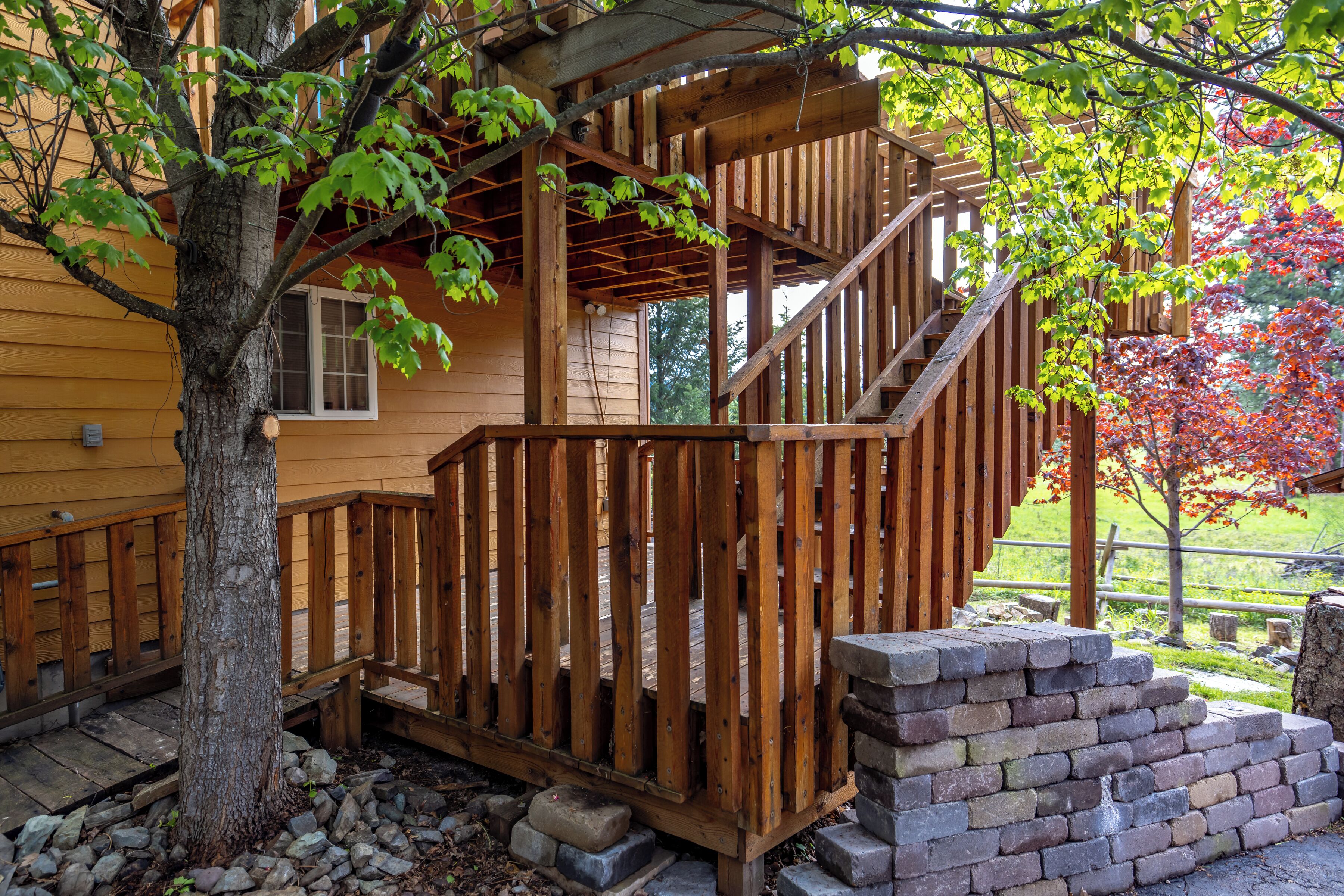 A wooden deck with a staircase leading to a porch, surrounded by trees and a stone wall.