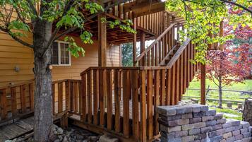 A wooden deck with a staircase leading to a porch, surrounded by trees and a stone wall.