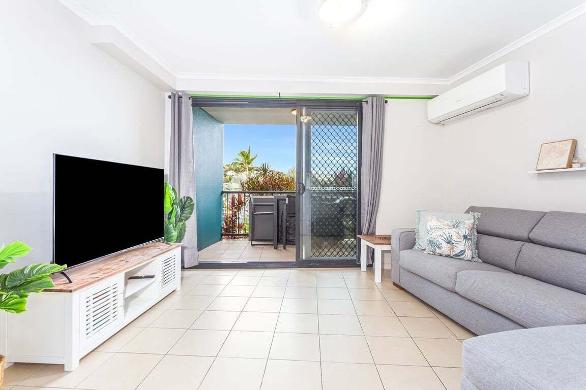 A modern living room with a flat-screen TV, a grey sofa, a small table, and a sliding glass door leading to a balcony with plants.