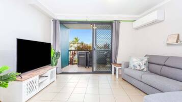 A modern living room with a flat-screen TV, a grey sofa, a small table, and a sliding glass door leading to a balcony with plants.