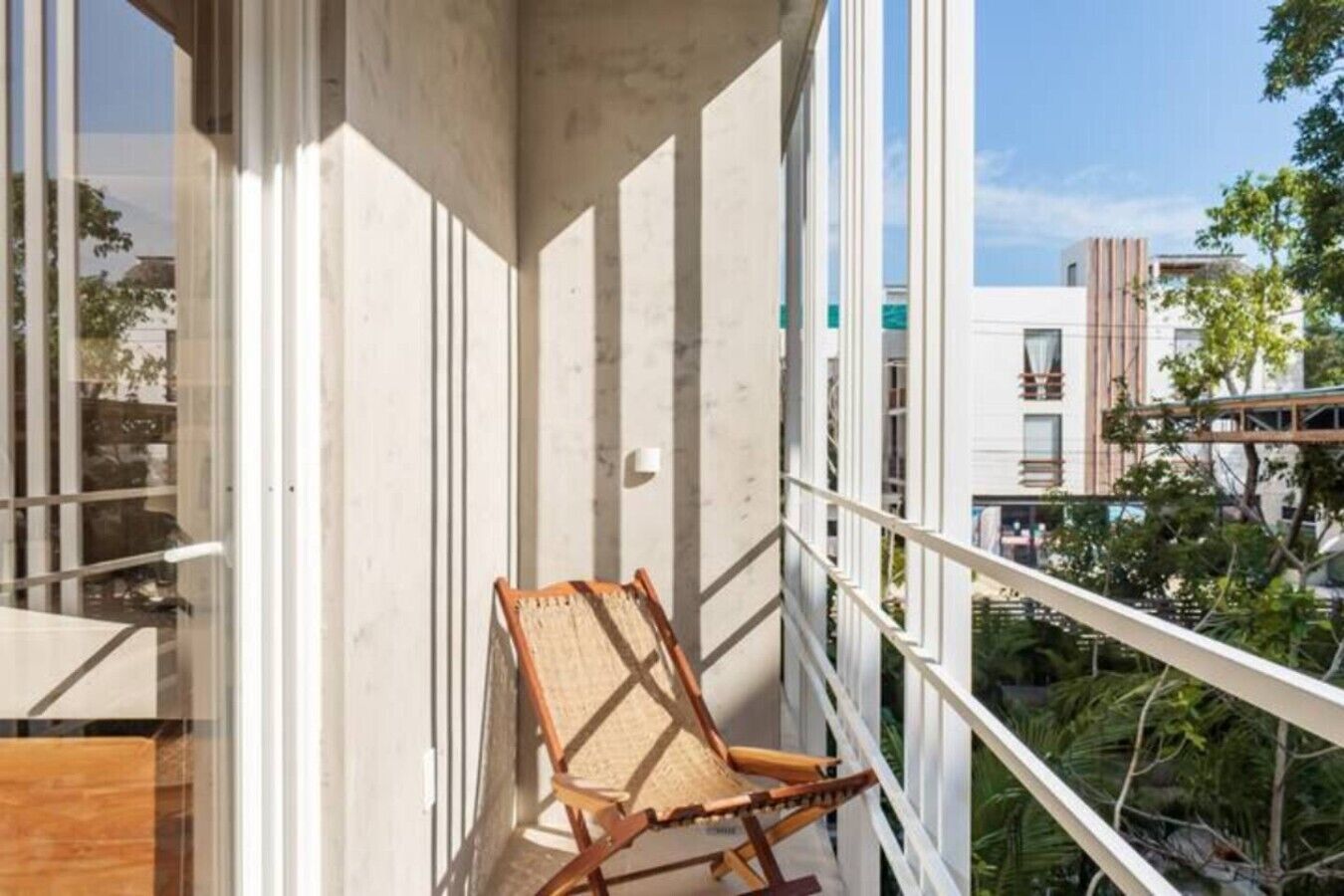 Un balcon avec une chaise en bois, de grandes fenêtres et une vue sur les bâtiments et la verdure.