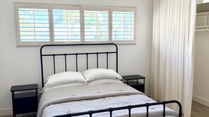 Un lit avec un cadre en métal, deux tables de chevet, un luminaire suspendu et une fenêtre avec des stores.