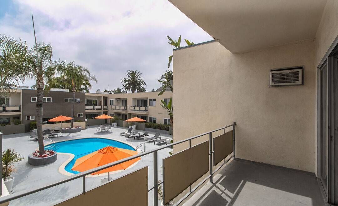 Uma área de piscina com espreguiçadeiras e guarda-sóis, uma varanda com corrimão, e um edifício com várias janelas e varandas ao fundo.