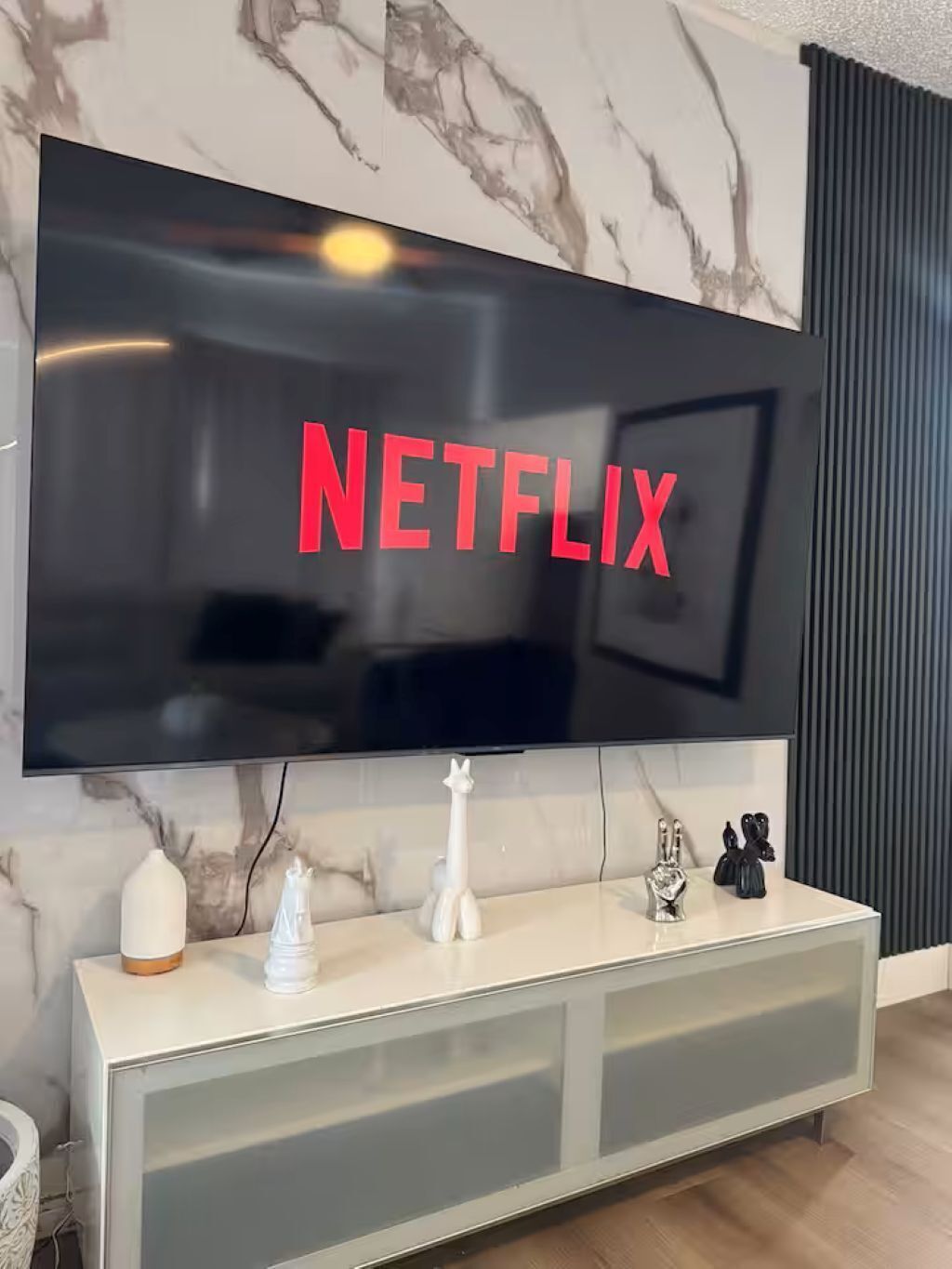Ein modernes Wohnzimmer mit einem großen Flachbildfernseher an einer Marmorwand, einem weißen Konsolentisch mit Dekoration und einer dunkelgrauen Akustikplatte rechts.