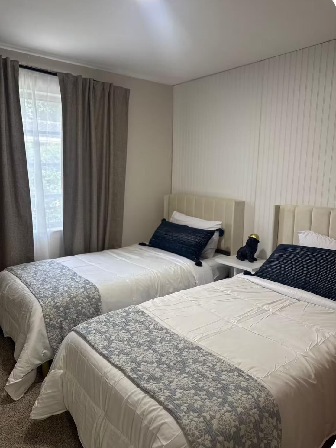Habitación de hotel con dos camas, una mesita de noche, una lámpara y una ventana con cortinas.