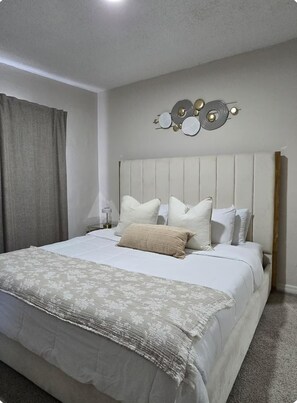 Un dormitorio con cama, almohadas, cabecera, decoración en la pared y cortina.