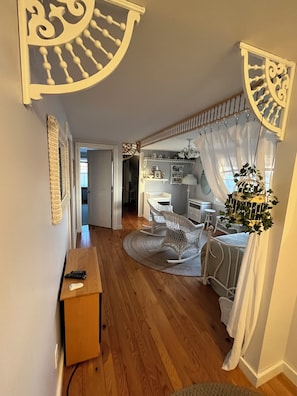 Ein Zimmer mit Holzboden, einem Schreibtisch mit Tastatur und Maus, einem Schaukelstuhl, einem kleinen Tisch mit Pflanze und einem Fenster mit Vorhängen.