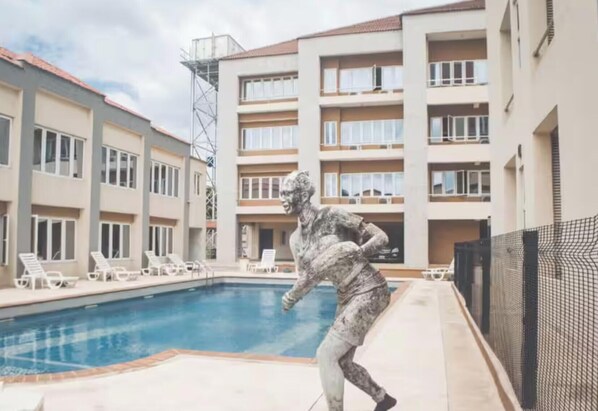 A swimming pool with lounge chairs, a statue, and a multi-story building with balconies. - Two-bed apt, ensuite rooms & close to amenities (Ikeja)