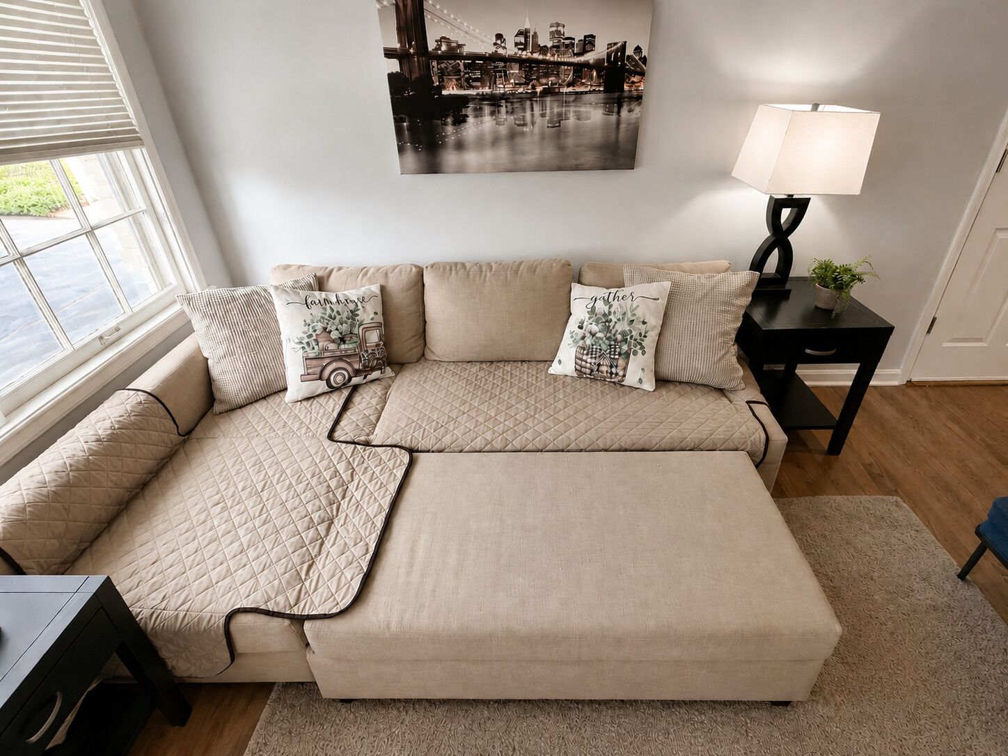 A modern living room with a large beige sectional sofa, a black side table with a lamp, and a cityscape painting on the wall.