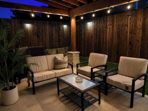Eine Terrasse im Freien mit Holzlattenzaun, Kamin aus Stein und einer Sitzecke mit einer Couch und zwei Stühlen.