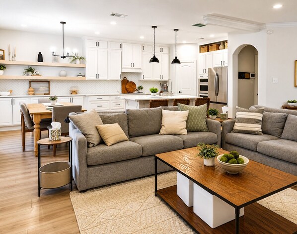 Eine moderne Küche mit weißen Schränken, ein Essbereich mit Holztisch und -stühlen sowie ein Wohnzimmer mit grauem Ecksofa und Couchtisch.