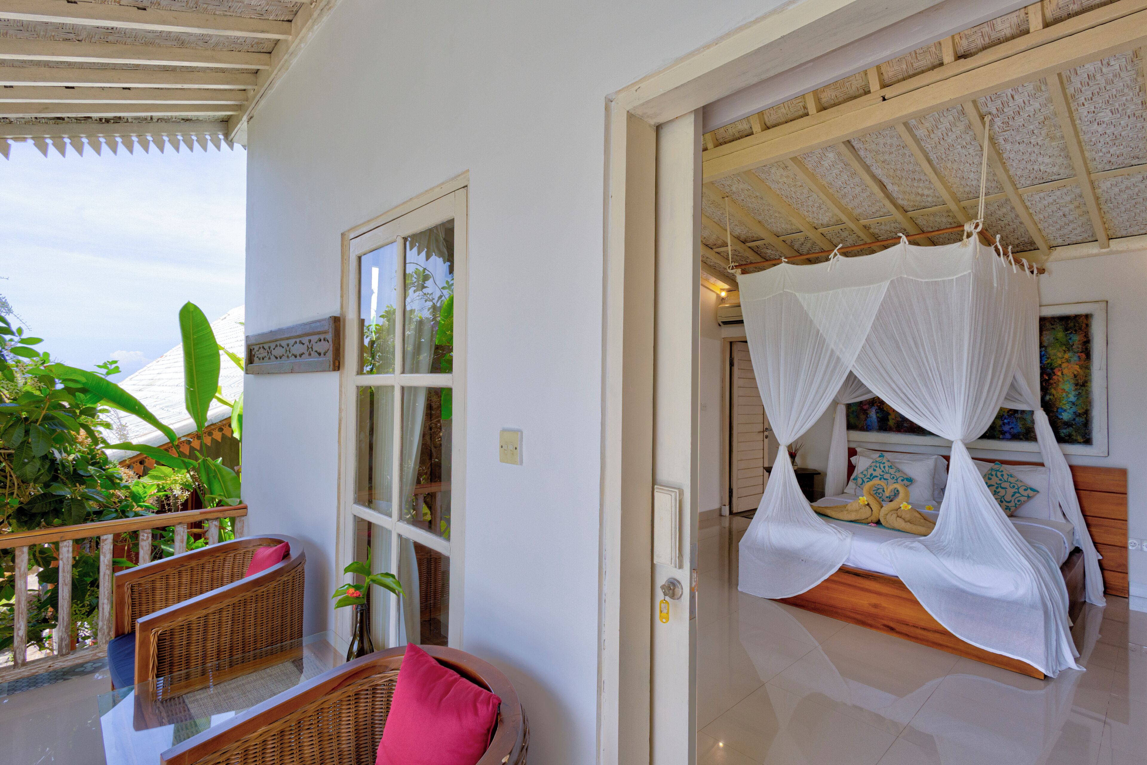 A room with a bed under a canopy, a wicker chair, and a view of greenery outside.