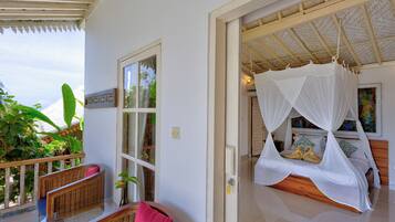 A room with a bed under a canopy, a wicker chair, and a view of greenery outside.
