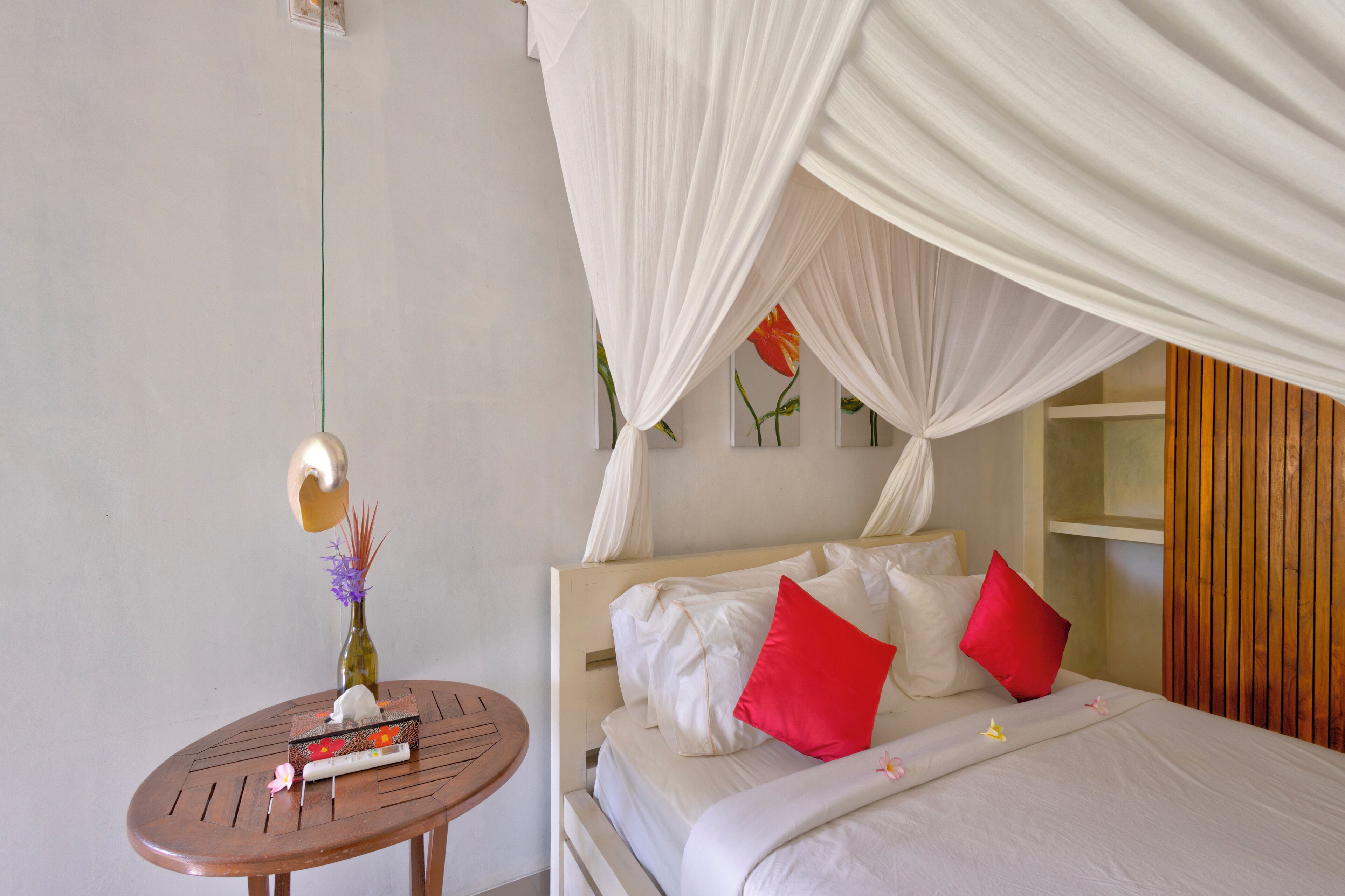 Ein Bett mit weißer Bettwäsche und roten Kissen, ein Holztisch mit einer Vase und Büchern, und eine Wand mit einem floralen Gemälde.