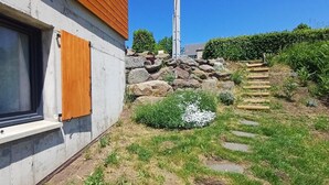 Ett hus med ett fönster, träpersienner, en betongvägg, en stenlagd gång och en trädgård med stenar och växter.
