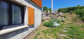 Ein Haus mit Fenster, Holzhörnern, einer Betonmauer, einem Kiesweg und einem Garten mit Steinen und Pflanzen.