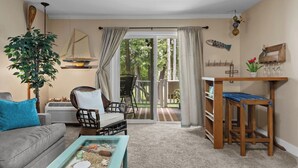 A living room with a sofa, chairs, a coffee table with a glass top, a wooden side table, a potted plant, a sailboat decoration, and a view through a glass door to an outdoor area. - Victorian Inn #107 | King Bed + Queen Sleeper Sofa (Elkhart Lake)