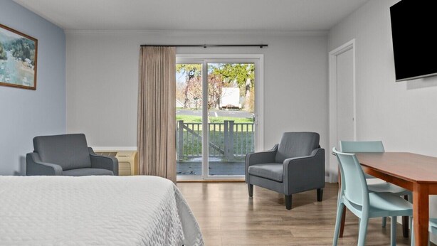 A hotel room with a bed, two armchairs, a dining table, a TV, and a sliding glass door leading to an outdoor area. - Victorian Inn #117 | King Bed (Elkhart Lake)