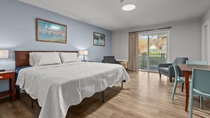 A hotel room with a bed, two bedside tables, a desk with chairs, a balcony with a view, and two paintings on the wall. - Victorian Inn #244 | King Bed (Elkhart Lake)