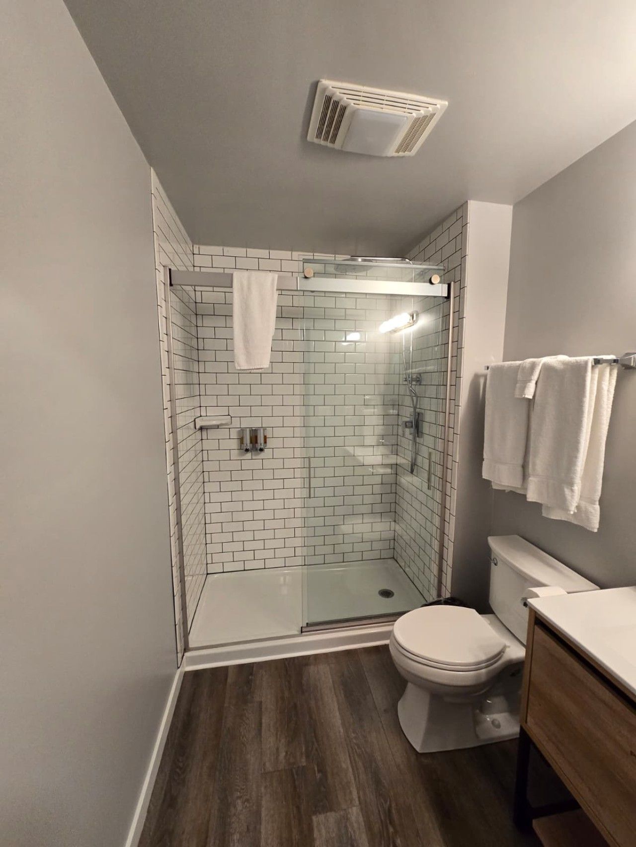 Une salle de bain moderne avec une douche à parois de verre, des carreaux blancs, des toilettes et un meuble-lavabo en bois.