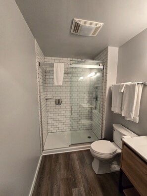 Une salle de bain moderne avec une douche à parois de verre, des carreaux blancs, des toilettes et un meuble-lavabo en bois.