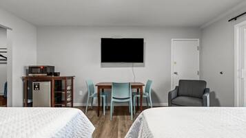 A room with two beds, a TV mounted on the wall, a dining table with chairs, a refrigerator, and a sofa.