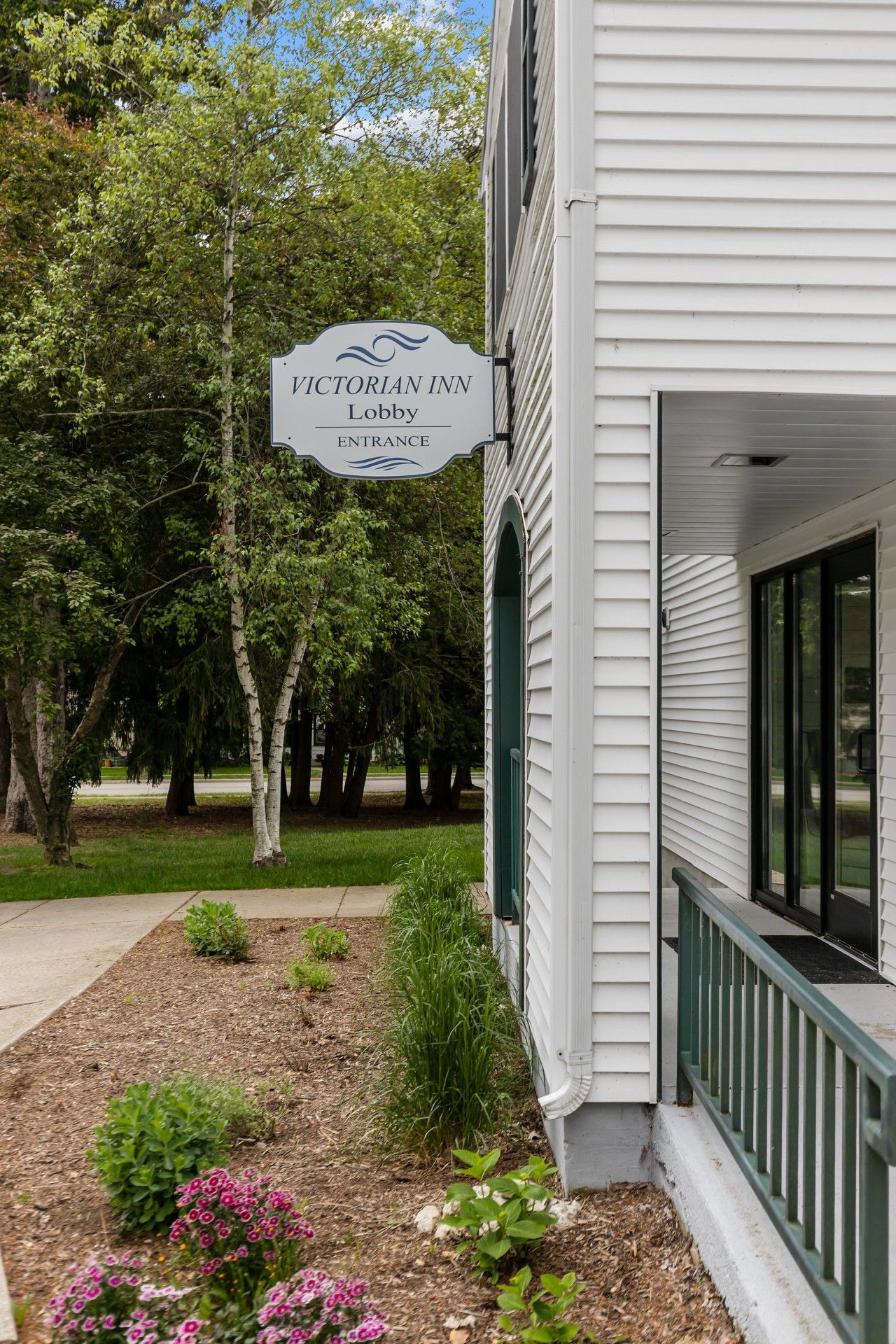 A Victorian Inn Lobby entrance sign, a white building with a green porch railing, and a landscaped garden with various plants.