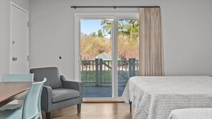 Un dormitorio con cama, una silla, una mesa y un ventanal corredero que da a un espacio exterior con vistas a árboles y una valla.