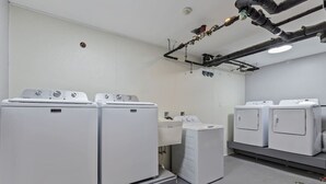 Una sala de lavandería con varias lavadoras y secadoras, un panel de control y tuberías aéreas.