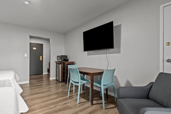 A hotel room with a wooden floor, a TV mounted on the wall, a dining table with chairs, a sofa, and a small kitchenette with a refrigerator and microwave. - Victorian Inn #239 | 2 Queen Beds (Elkhart Lake)