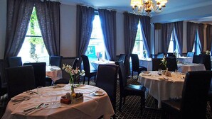 En restaurant med runde borde, stole, en lysekrone og store vinduer med gardiner.