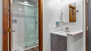 A bathroom with a glass shower enclosure, a sink with a marble countertop, and a vanity with a mirror and lights above. - Victorian Inn #246 | 2 Queen Beds (Elkhart Lake)