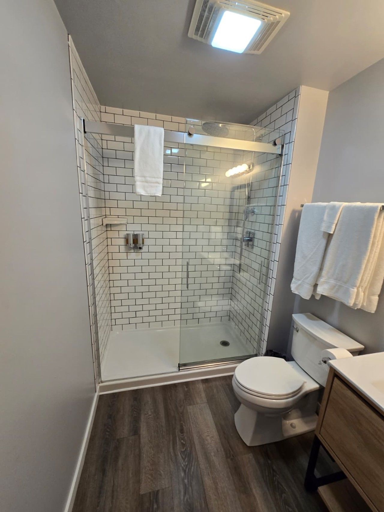 Une salle de bain moderne avec une douche à parois de verre, des murs recouverts de carreaux blancs, des toilettes et un meuble-lavabo en bois.