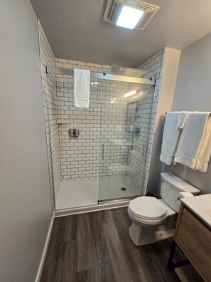 Un baño moderno con cabina de ducha de vidrio, paredes con azulejos blancos, inodoro y tocador de madera.