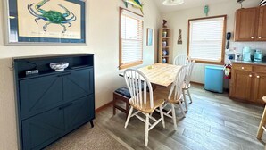 A dining area with a table and chairs, a blue cabinet, a framed artwork, and a kitchen in the background. - Beach Dreamer with Ocean Views, 120 Steps to the Beach • Buxton, NC, OBX (Buxton)