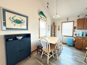A dining area with a wooden table and white chairs, a black cabinet, and a framed picture of a blue crab on the wall.