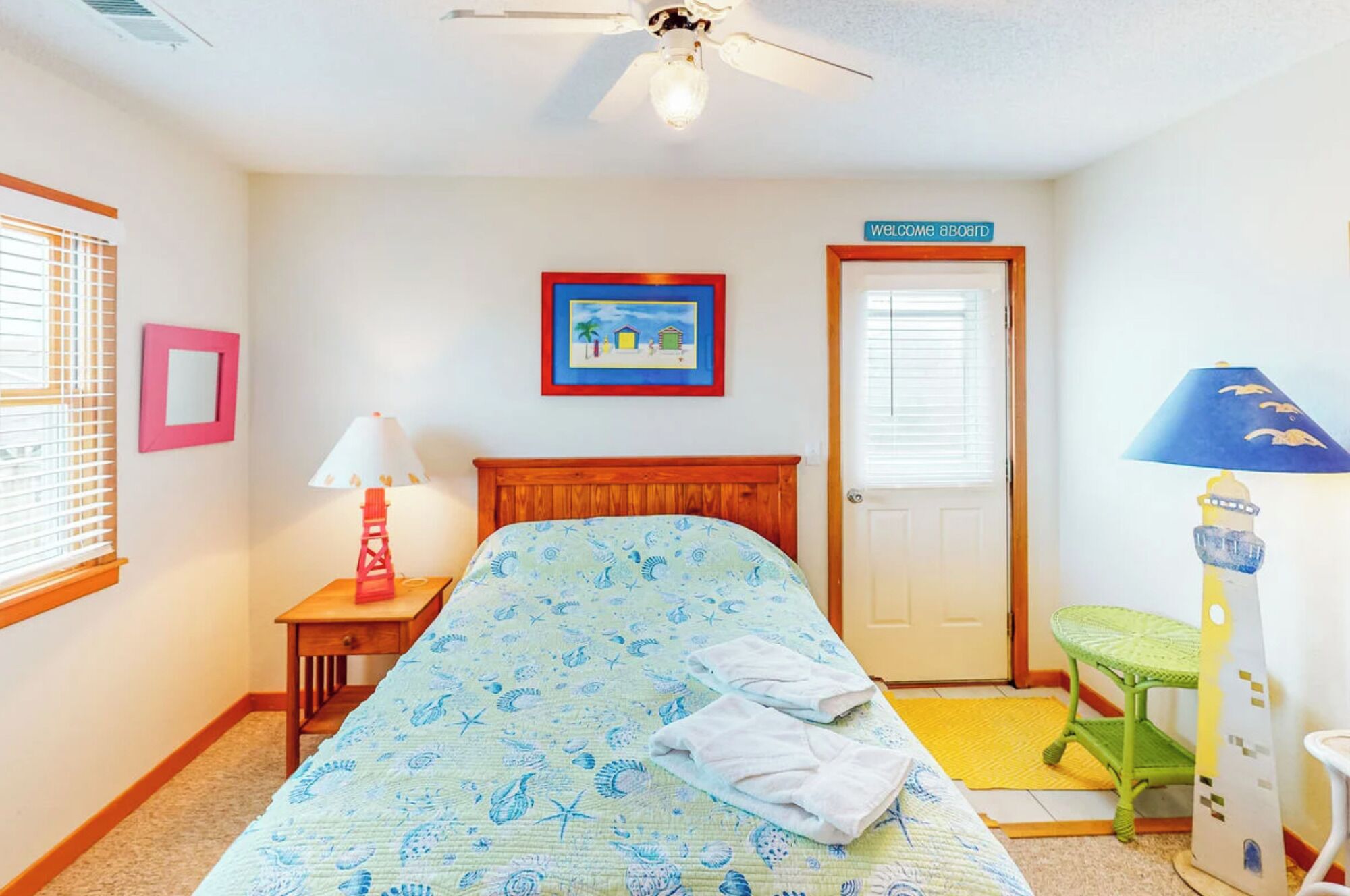 A bedroom with a bed, a nightstand, a lamp, a framed picture, a door with a sign, and a ceiling fan.