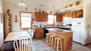 A kitchen with wooden cabinets, a central island with stools, a dining table with chairs, and pendant lights. - Beach Dreamer with Ocean Views, 120 Steps to the Beach • Buxton, NC, OBX (Buxton)