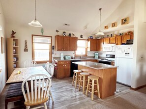 A kitchen with wooden cabinets, a central island with stools, a dining table with chairs, and pendant lights.