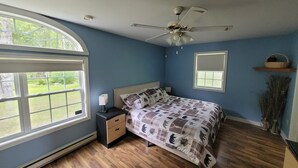 Un dormitorio con cama, mesita de noche, ventilador de techo, ventana y un estante pequeño.