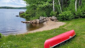 Un canoa rojo en la orilla cubierta de hierba junto a un lago, con árboles y rocas al fondo.