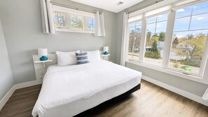 Un dormitorio con una cama grande, dos mesitas de noche, una ventana con cortinas y vistas a una zona residencial.