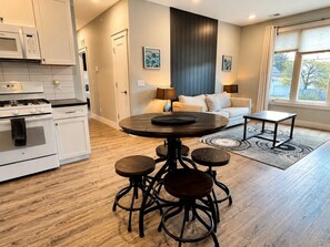 A modern kitchen with a central island, a dining area with stools, a sofa, and a window with curtains. - The Haven 10 | 2BR | Rooftop Patio |Downtown (Traverse City)