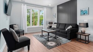 Ein modernes Wohnzimmer mit Sofa, Couchtisch und Sesseln, einem an der Wand befestigten Fernseher sowie einem großen Fenster mit Vorhängen.