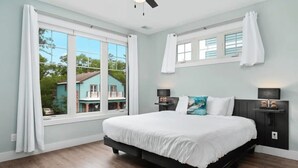Ein Schlafzimmer mit einem großen Bett, zwei Nachttischen, einem Deckenventilator und Blick auf ein Haus durch ein Fenster.