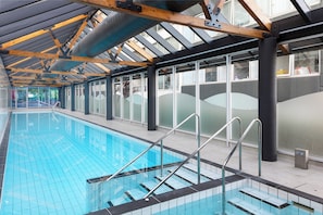 Piscina interna com paredes de vidro transparente, guarda-corpos de metal e teto alto com vigas de madeira.
