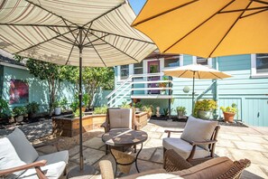 Eine Terrasse im Freien mit Korbmöbeln, einem runden Tisch und zwei Sonnenschirmen, die Schatten spenden.
