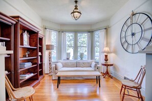 Ein Zimmer mit einer Holzbibliothek, einem weißen Sofa, einem Holzstuhl, einem runden Tisch und einer Wanduhr.