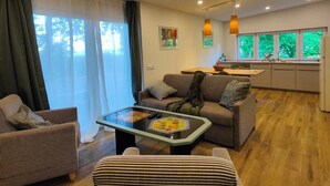 Ett modernt vardagsrum med en soffa, fåtöljer, ett soffbord i glas och en köksdel med utsikt genom ett fönster.